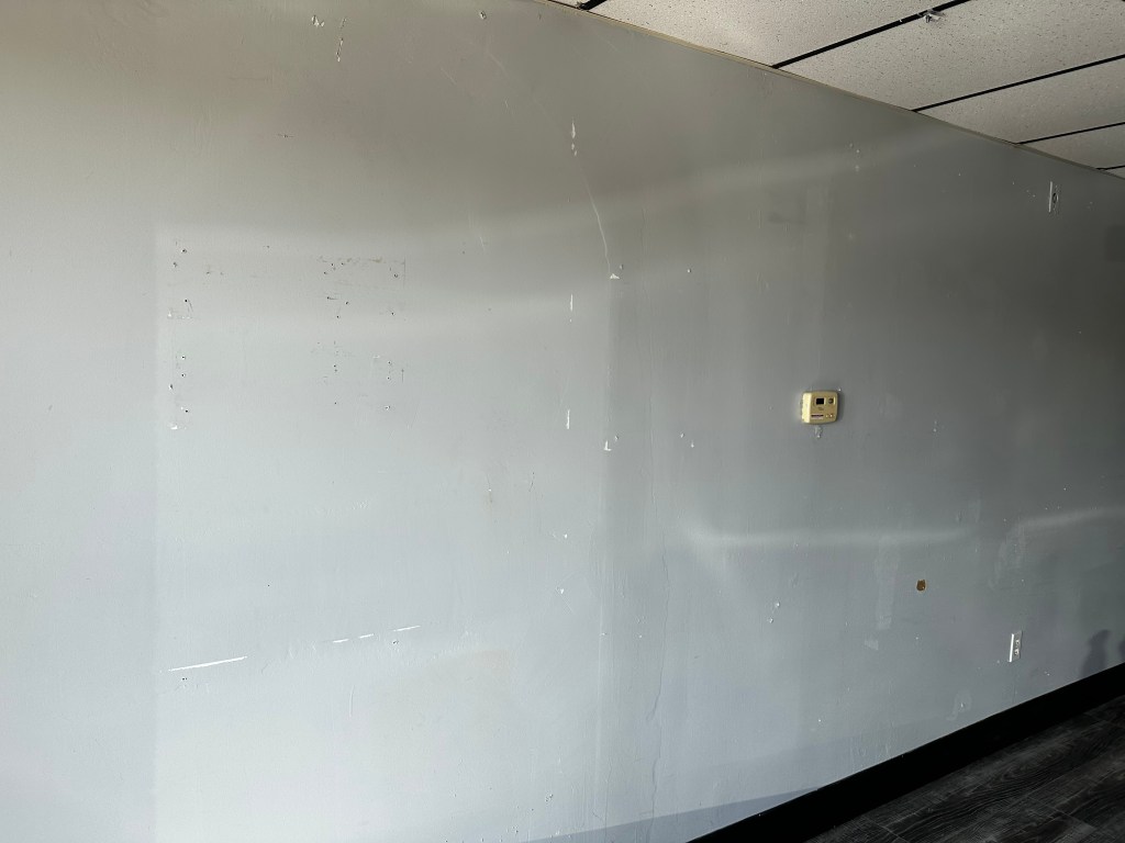 A plain gray wall with minor blemishes and a thermostat mounted on it, in a sparsely decorated room.
