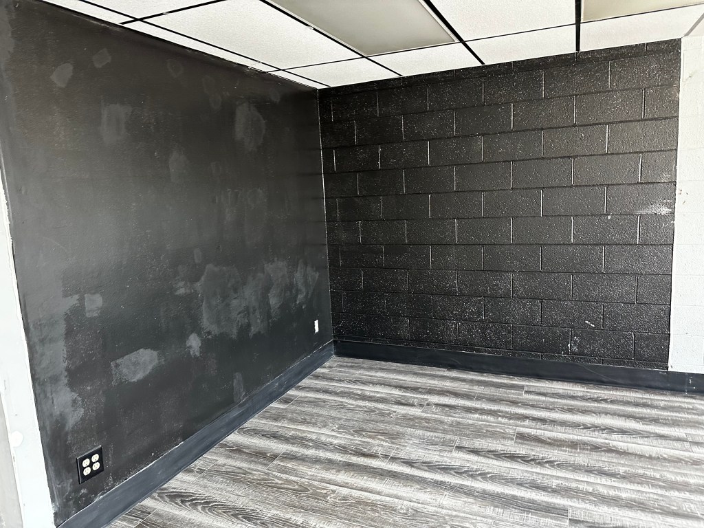 A partially empty room featuring a black wall on one side and a textured black block wall on the other, with light-colored wooden flooring.
