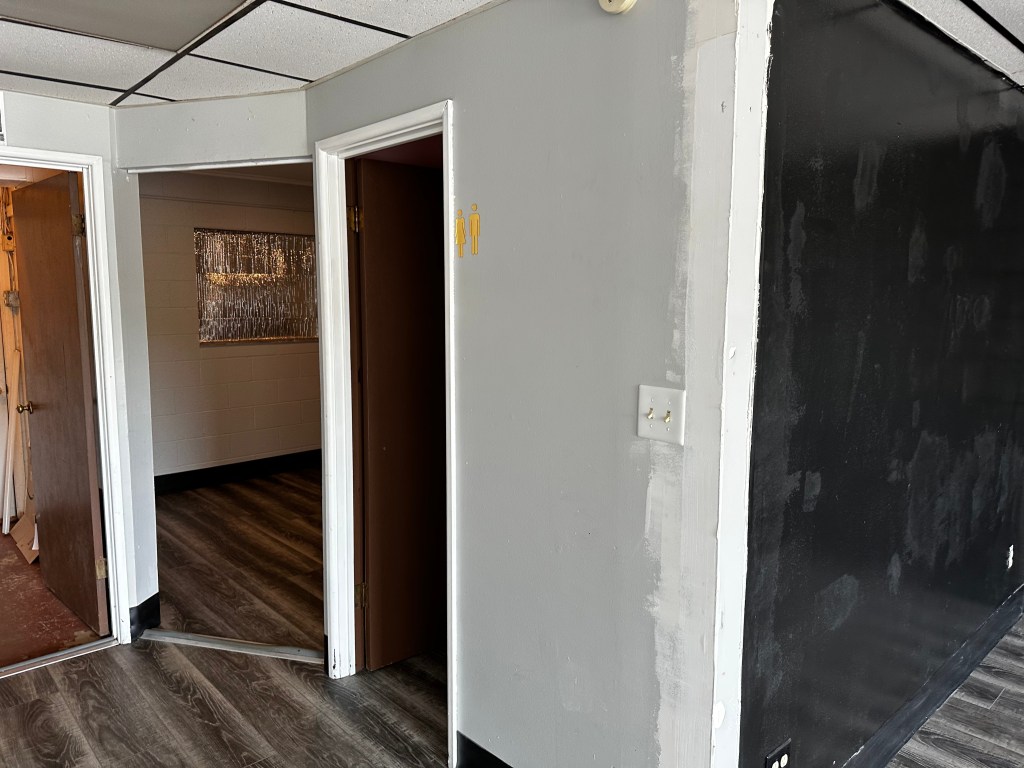 Interior view of a room featuring a doorway with a gray wall on one side and a black wall on the opposite side, wooden doors partially open, and a patterned window in the background.