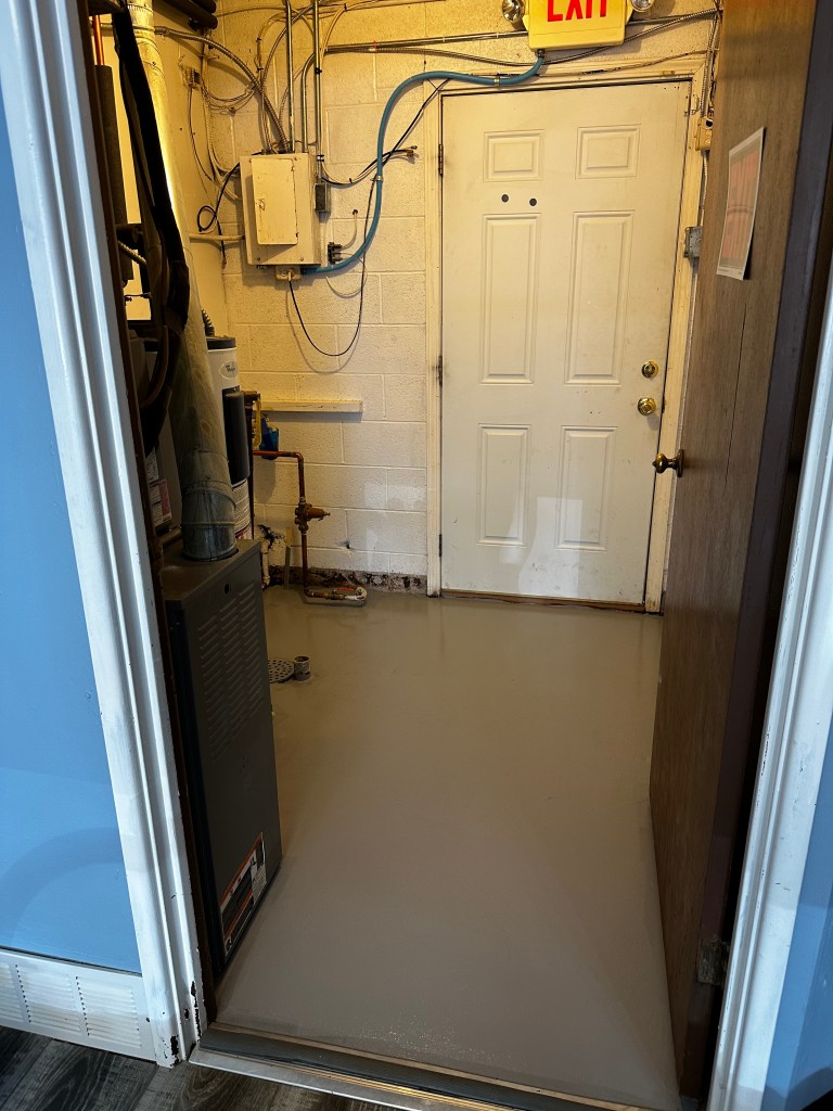 A view of an interior entrance with a partially open door, showing a freshly coated grey floor with visible plumbing and electrical fixtures along the walls.