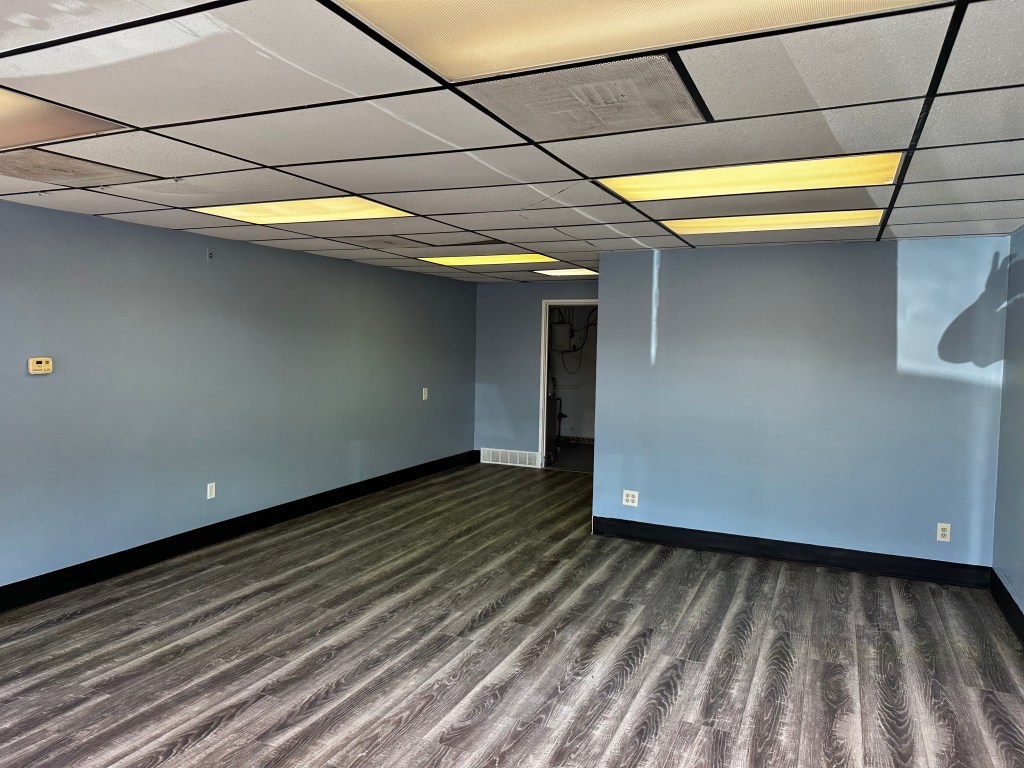 Empty room with light blue walls and grey wood flooring, featuring fluorescent ceiling lights and a doorway at the back.