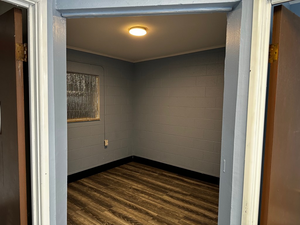 Interior view of a small room with gray walls, a wooden floor, and a ceiling light. A window is visible in the background, and there are two doorways on either side.